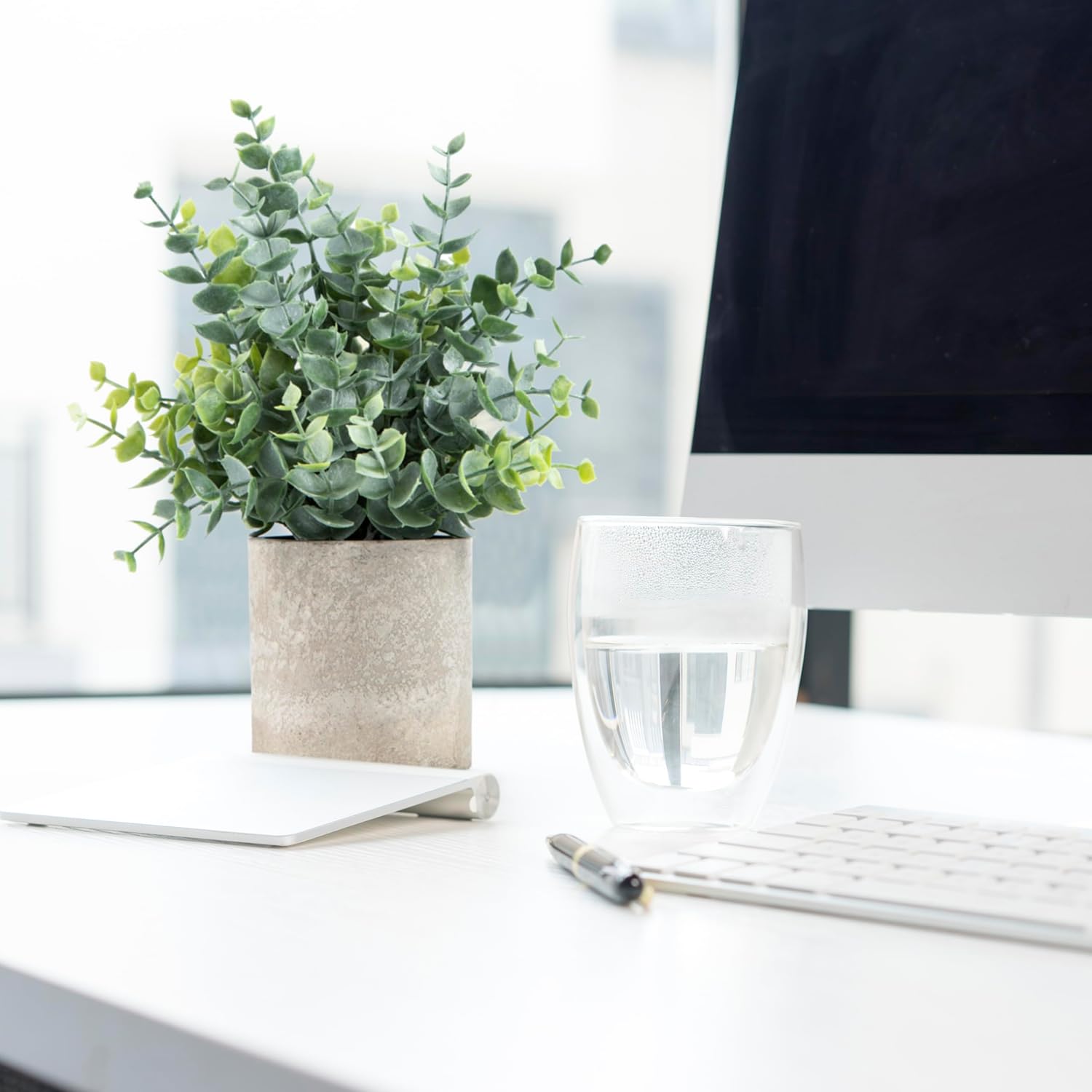 Decorative artificial eucalyptus plants in a modern pot on a desk next to a glass of water and a notebook