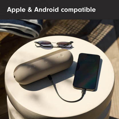 Beats Pill Bluetooth Speaker in beige on a table with sunglasses and a phone connected